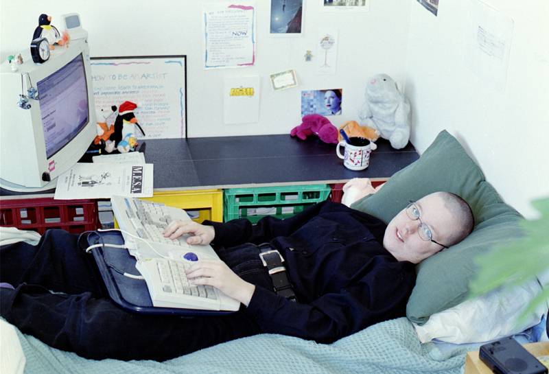 1999: a person lying in bed with a keyboard on their lap, beside a desk holding a monitor and several soft toys