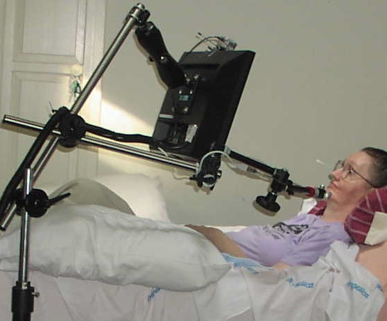 A person lying in bed with a large monitor held in front of them on a jointed metal arm attached to a floor stand