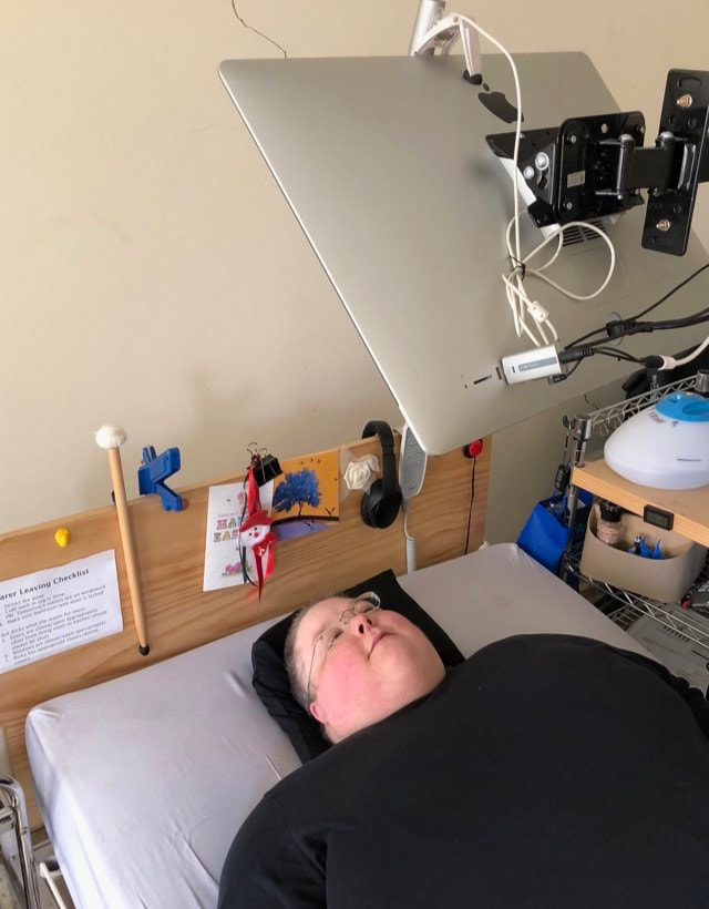 A short-haired white woman lies in bed, a computer mounted over her hair so she can see it lying down